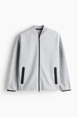 Campera bomber deportiva gris melange, de corte estándar, con cuello elevado corto, cierre bidireccional y mangas raglán. Posee bolsillos delanteros con cierre y costuras selladas, además de elástico fino en puños y bajo.