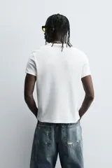 Camiseta de corte relajado, color gris claro, confeccionada en tejido con estructura waffle. Presenta cuello redondo y manga corta, con costuras decorativas en contraste color dorado que recorren los laterales y los hombros.