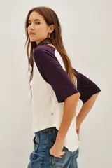 Remera estilo beisbolera con mangas raglán en color violeta oscuro y cuerpo en tono crema. Presenta una estampa frontal con texto en color negro.