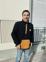 Buzo de hombre de polar negro con cierre parcial color naranja, bolsillo de parche color naranja en la parte inferior y cuello alto.