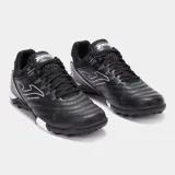Championes de fútbol Joma modelo Maxima, diseñados para césped sintético. Presentan un diseño en color negro con detalles en blanco, confeccionados con materiales sintéticos de alta resistencia y sistema de ventilación integrado.