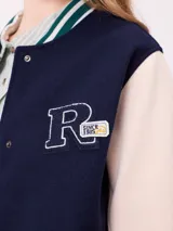 Campera estilo varsity con cuerpo en color marrón y mangas en tono beige. Presenta cierre frontal con botones a presión, cuello, puños y ruedo con franjas tejidas en verde y blanco. Incluye un parche con la letra R en el pecho y detalles bordados en las mangas.