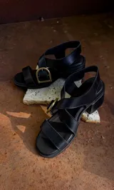 Sandalias de cuero color marrón con tiras anchas cruzadas y hebilla metálica decorativa en forma de estribo. Presentan taco cuadrado de altura media y suela de goma.