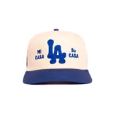 Gorro trucker color beige con visera y detalles bordados en azul que incluyen las letras "LA" y el texto "Mi Casa Su Casa".