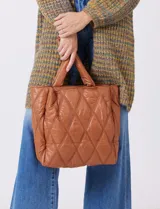 Cartera de eco cuero color marrón, con diseño capitoneado en rombos de gran relieve. Cuenta con un compartimento principal con cierre, amplios bolsillos laterales con botones imantados, asa de mano fija y correa larga regulable y removible. El forro interno es textil a tono con compartimentos organizadores.