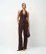 Chaleco de sastrería color marrón, con diseño de frente única y cuello halter. Presenta pinzas laterales que definen la silueta, bolsillos decorativos y ruedo levemente redondeado. En la espalda, cuenta con una cinta ajustable con ojal metálico y tiras largas.