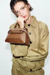 Bolso de mano tipo city color marrón, con diseño estructurado, costuras a la vista en color blanco y detalles de hebillas metálicas decorativas en la parte frontal. Cuenta con asa de mano superior, correa ajustable para usar como bandolera y cierre mediante solapa con imán.