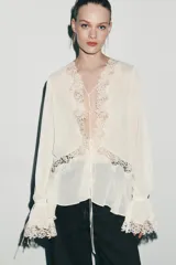 Blusa de escote en pico con manga larga terminada en volantes. Presenta aplicaciones de encaje a tono, cierre frontal con lazadas y bajo asimétrico.