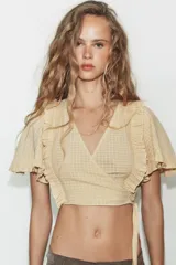 Top crop de escote en pico y manga corta con volados. Presenta un diseño cruzado que se ajusta mediante una lazada lateral y cuenta con una textura de cuadrícula sutil en toda la prenda.