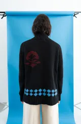 Cardigan de punto color marrón con cierre frontal, cuello alto y bolsillos aplicados. Presenta diseño tejido localizado con detalles en violeta y negro en el pecho.