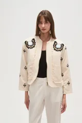 Campera estilo bomber color beige claro, con cuello redondo, cierre frontal y detalles de volados en los bordes. Presenta bordados florales en negro y blanco en la zona del pecho y bordados de tréboles negros en las mangas.