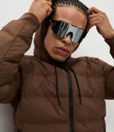 Campera puffer deportiva de color marrón, con capucha ajustable mediante cordones, cierre frontal de cremallera y puños elásticos. Diseño acolchado sin costuras visibles.
