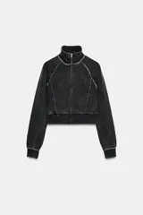 Sudadera corta negra con efecto lavado, cuello alto y manga larga. Presenta costuras marcadas a contraste en color blanco y cierre frontal con cremallera metálica.