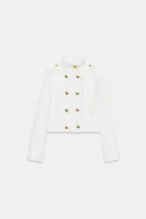 Chaqueta corta blanca de estilo militar, con cuello subido, manga larga y trabillas desflecadas en los hombros. Cuenta con cierre frontal cruzado decorado con botones metálicos dorados.