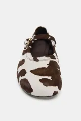 Zapato plano tipo bailarina con acabado de piel y estampado animal print de manchas marrones y blancas. Presenta una tira ajustable con hebilla metálica sobre el empeine y punta redonda.