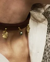 Gargantilla tipo choker de gamuza con dijes colgantes artesanales de metal y perlas naturales. Cuenta con tiras largas para atar y ajustar en la parte posterior.