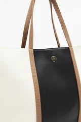 Cartera tipo tote bag en símil cuero, con diseño en bloques de color negro y blanco, y detalles en marrón. Tiene asas largas para llevar al hombro y un cierre magnético.