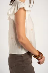 Blusa blanca sin mangas con escote en V, cierre frontal con botones y mangas con volados. Presenta pequeños bordados sutiles distribuidos por toda la prenda.