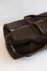 Bolso de viaje tipo weekender de cuero vacuno color marrón chocolate, con doble asa de mano y correa bandolera ajustable y extraíble. Tiene cierre superior con cremallera y múltiples bolsillos internos.