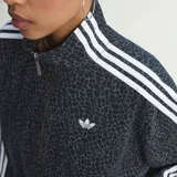 Campera deportiva Adidas Originals modelo Firebird con cierre frontal completo, cuello alto y puños elásticos. Presenta un estampado integral de patrón orgánico en tonos oscuros, con las icónicas tres tiras blancas en los hombros y mangas, y el logo del trifolio bordado en el pecho.