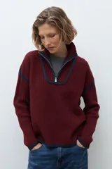 Sweater de punto color crudo con cuello alto y cierre frontal parcial. Presenta detalles de vivos en color marrón en el cuello, hombros y ruedo, con diseño de manga larga.