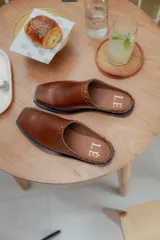 Mules de cuero color suela, con diseño abierto y tira frontal decorada con tachas metálicas circulares. Presentan punta cuadrada y suela plana.
