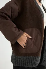 Saco tipo cardigan de punto gris con diseño bicolor, mangas largas acolchadas negras y bolsillos frontales también acolchados en color negro. Presenta cierre frontal con botones y terminaciones en tejido acanalado.