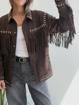 Chaqueta de efecto gamuza en color chocolate, con diseño de flecos en el pecho y mangas. Presenta detalles de tachas metálicas plateadas a lo largo de los hombros, solapas y bolsillos. Cuenta con cierre frontal de botones y forro interior.