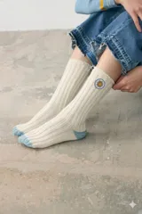 Medias de tejido acanalado en color crudo, con puntera y talón en color celeste. Presentan un parche bordado circular con el diseño del sol de la bandera de Uruguay en la parte superior.