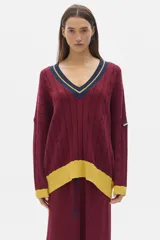 Buzo tejido de punto con diseño oversized, cuello en V y mangas largas. Presenta un patrón de trenzas verticales y detalles en contraste de color amarillo en el ruedo y los puños, además de un borde azul marino en el cuello.