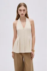 Blusa de algodón color beige con escote en V, cuello halter y cierre frontal con botones. Presenta un diseño ajustado en el torso con pinzas y pliegues en la parte inferior que generan una caída ligeramente acampanada.