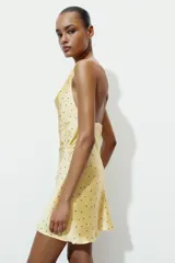 Vestido corto de satén con estampado de lunares, escote fluido tipo cowl neck y tirantes finos que se cruzan en la espalda con una aplicación metálica circular.