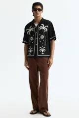 Camisa de manga corta de H&M, con corte relajado, cuello abierto estilo resort, tapeta frontal con botones y bajo recto. Diseño estampado en color blanco sobre base negra con motivos de palmeras, soles y estrellas.