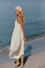Vestido largo satinado color crudo, con escote en pico adornado con lazada y tirantes finos ajustables. Presenta detalles de frunces y diseño fluido.