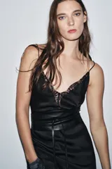 Cinturón tipo fajín ancho de piel sintética negra, con forma curva en la parte superior e inferior. Presenta detalles metálicos dorados en los laterales y se ajusta mediante lazos largos del mismo material.