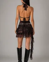 Vestido corto de dos piezas con estampado tipo tie-dye en tonos oscuros. El top es estilo halter con escote drapeado, mientras que la falda es de corte mini con volados en capas y una tira lateral larga con volados decorativos. Incluye un cinturón de cadena metálica con eslabones circulares.