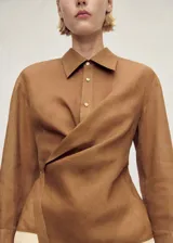 Camisa de ramio color tostado con cuello camisero, manga larga con puños abotonados y cierre frontal parcial con tres botones. Presenta un diseño cruzado en el frente y bajo asimétrico.