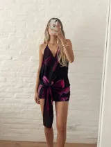 Vestido corto de escote halter con espalda descubierta, confeccionado en tela fluida con estampado floral en tonos violeta y fucsia sobre base negra. Presenta un diseño cruzado con un nudo frontal que crea un efecto drapeado.