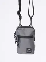 Bandolera de lona color gris con detalles en negro, cierre principal y bolsillo frontal con cierre. Incluye cordón decorativo en el lateral y correa ajustable con ganchos.