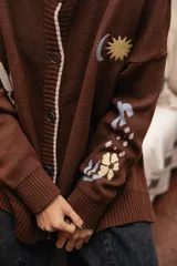 Saco tejido color chocolate con diseño inspirado en la granada, con flores y soles bordados. Presenta cierre frontal con botones, cuello redondo y puños acanalados.