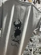 Remera gris de algodón con estampado frontal en negro del logo del videojuego "Hollow Knight", con tipografía decorativa y un emblema central.