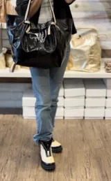 Bolso tipo tote de cuero negro con acabado brillante, diseño fruncido en los laterales y correas de hombro de tela tejida en tono beige.