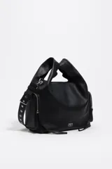Bolso bandolera de piel vacuna color negro, con estructura blanda y forma hobo o de media luna. Presenta dos bolsillos frontales con cierre metálico plateado y un asa de hombro ancha y ajustable con hebillas y ojales metálicos. El logo de Bimba y Lola está estampado en la parte frontal inferior.