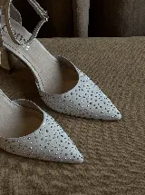 Zapato de tacón con punta fina, confeccionado en tela con aplicación de strass en la capellada y detalles en cuero en el talón, taco y pulsera. Cuenta con cierre de hebilla al tobillo y taco de altura media.