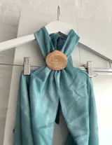 Top halter de color celeste/turquesa, con efecto drapeado y un accesorio tipo concha marina como broche central. El diseño es holgado y se ajusta en la espalda.