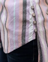Camisa sin mangas de corte recto, con cuello clásico y escote en V. Presenta un diseño de rayas verticales en tonos lila, rosa, blanco y gris, con dos bolsillos frontales aplicados con estampado en chevron. Cuenta con cierre central de botones.