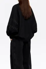 Sudadera negra de corte oversize, confeccionada en algodón, con escote redondo y mangas largas tipo ranglán. Presenta detalles de remaches metálicos distribuidos a lo largo de las costuras de las mangas.