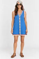 Vestido corto celeste de punto con escote en V, sin mangas, abotonado al frente con botones de nácar y detalles de ribetes en rojo y celeste en el escote y la botonera. El ruedo y las sisas presentan terminaciones en encaje beige.