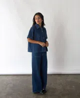 Camisa de denim azul de algodón reciclado, corte amplio, con cuello clásico, manga corta, cierre frontal con botones y dos bolsillos de parche en el pecho.