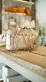 Cartera tipo bolso de cuero con acabado texturizado en tono beige y blanco, decorada con flecos largos y tachas metálicas circulares en la parte superior. Cuenta con doble correa de mano y cierre superior.